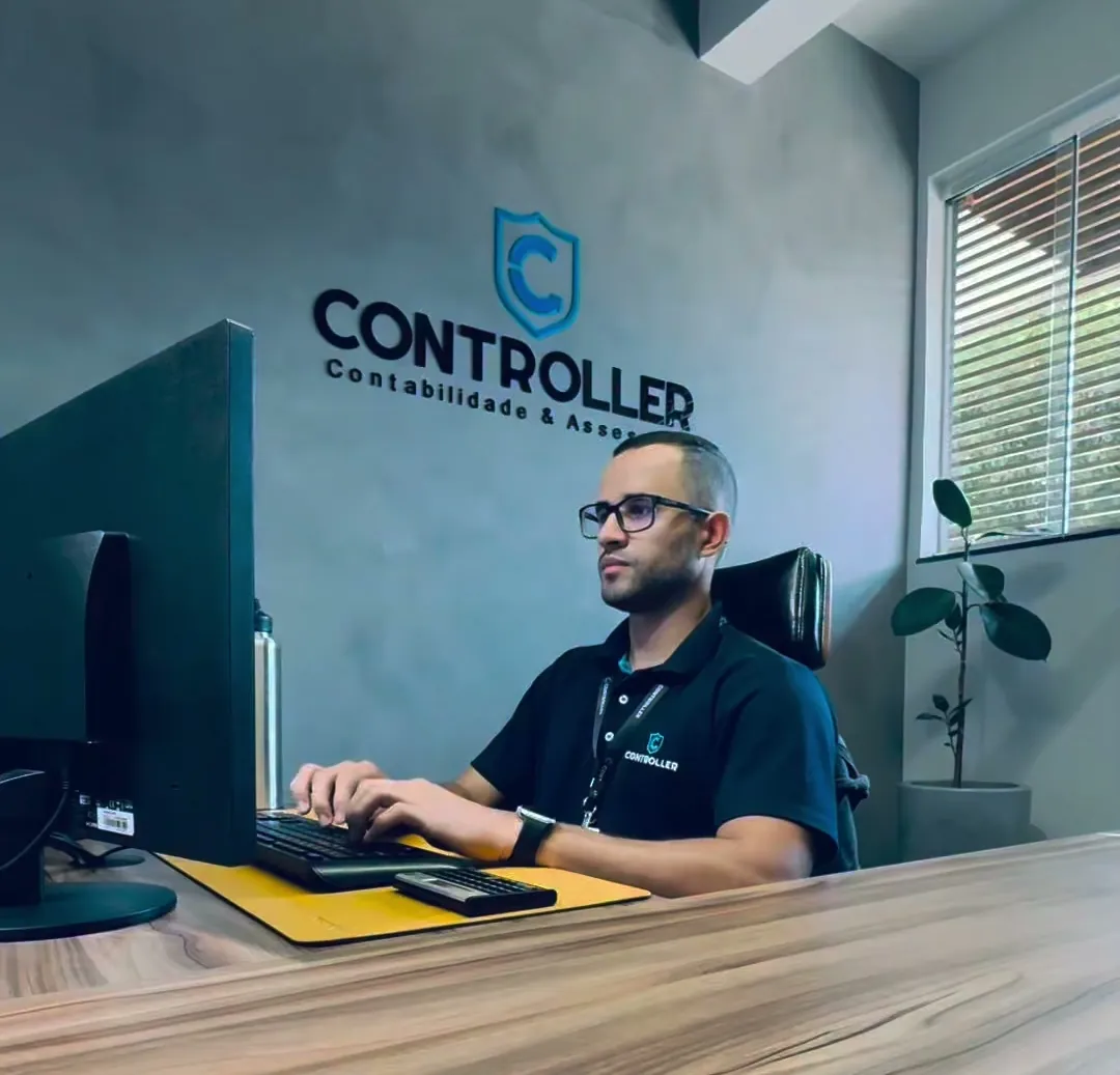 O contador Vitor Rodrigues Dias trabalhando no computador, na sala do escritório, com o logo da Controller na parede.